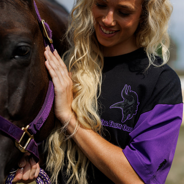 Model wearing the Two-Tone Lax Tee in black and purple beside a horse, close view