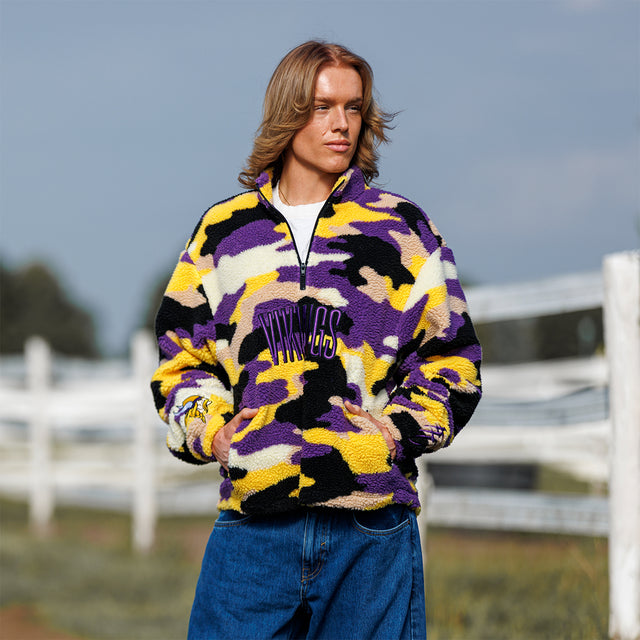 Model wearing the Sherpa 1/4 Zip Pullover in purple and gold camo, front view by a fence